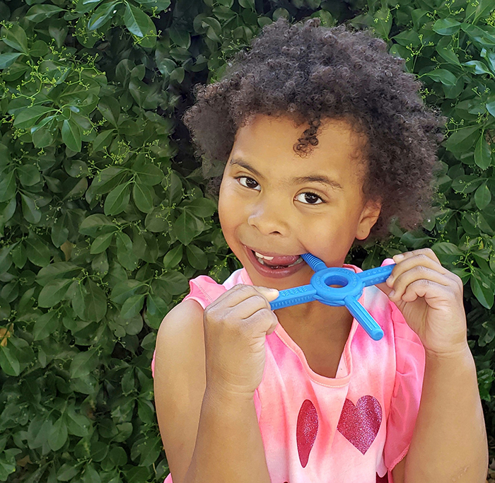 A girl chews on the royal blue Tetra-Bite handheld chew tool outside in front of green bushes