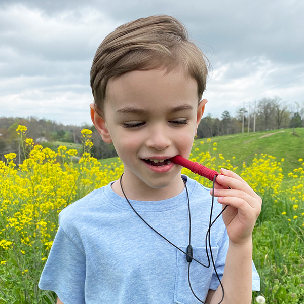 A boy chews on dark red Bite Saber chew necklace with his back molars outside in front of a meadow